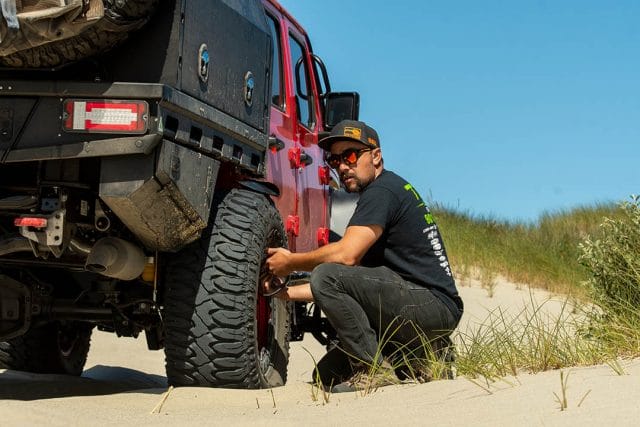 man checks the tire pressure on a red jeep gladiator on the beach
